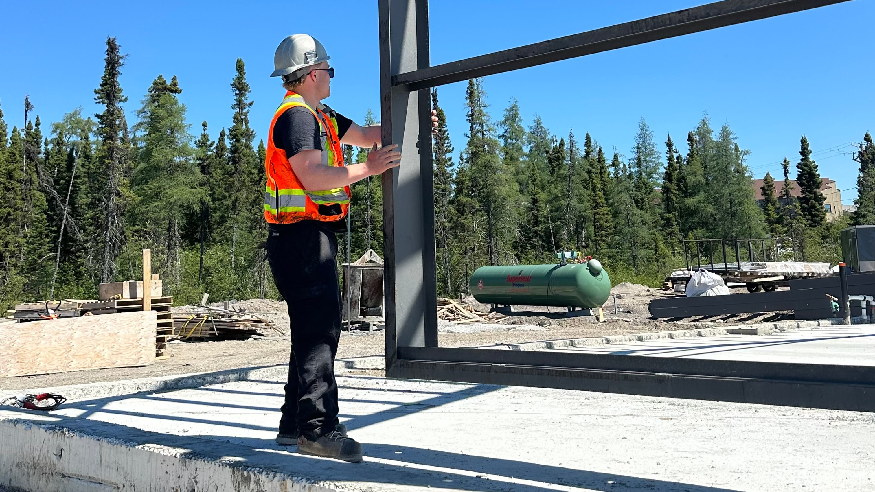 Person in safety gear working on a metal frame structure with trees and clear sky in the background